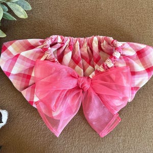 May include: A baby accessory with a pink and white plaid pattern, featuring a large, sheer pink bow. The item is gathered at the top, creating a ruffled effect. The overall design is soft and feminine.