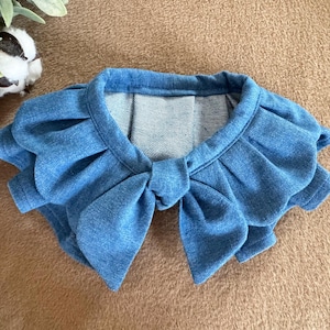 May include: A light blue denim bolero with a bow detail. The bolero features ruffled sleeves and a round neckline. The garment is made of a soft denim fabric and is designed to be worn as a top.