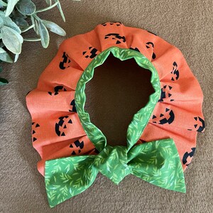 May include: Orange fabric collar with a green bow and a pumpkin face pattern. The collar is ruffled and has a green trim.
