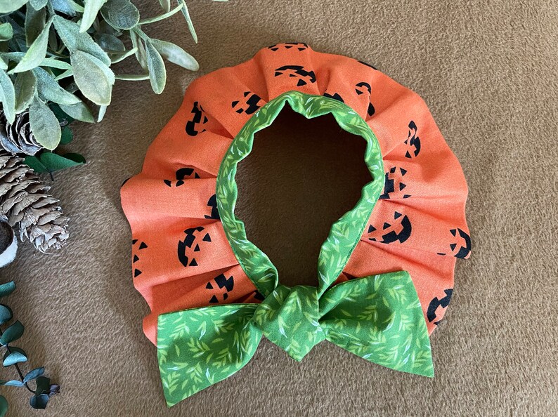 May include: Orange fabric collar with a green bow and a pumpkin face pattern. The collar is ruffled and has a green trim.