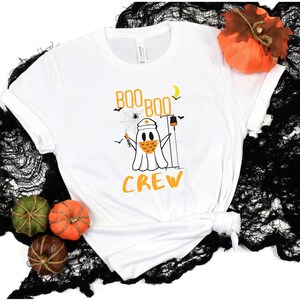 May include: White t-shirt with a ghost wearing a face mask and a nurse's hat. The ghost is holding a syringe and a thermometer. The text "Boo Boo Crew" is printed on the shirt.
