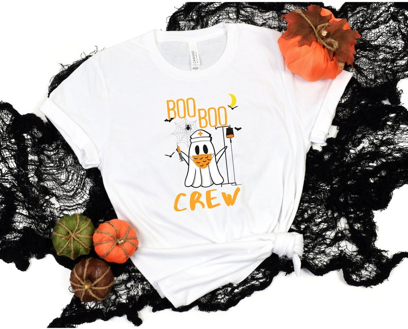 May include: White t-shirt with a ghost wearing a face mask and a nurse's hat. The ghost is holding a syringe and a thermometer. The text "Boo Boo Crew" is printed on the shirt.