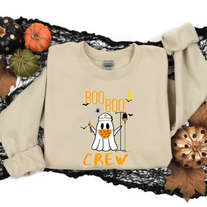 May include: A tan sweatshirt with a ghost wearing a face mask and a nurse's hat. The ghost is holding a syringe and a IV bag. The text "BOO BOO CREW" is printed on the sweatshirt.