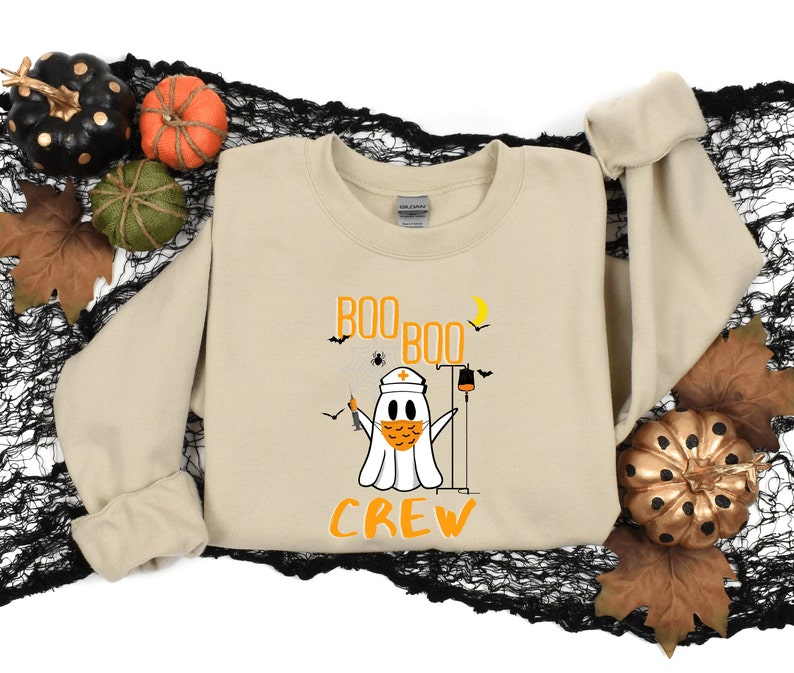 May include: A tan sweatshirt with a ghost wearing a face mask and a nurse's hat. The ghost is holding a syringe and a IV bag. The text "BOO BOO CREW" is printed on the sweatshirt.