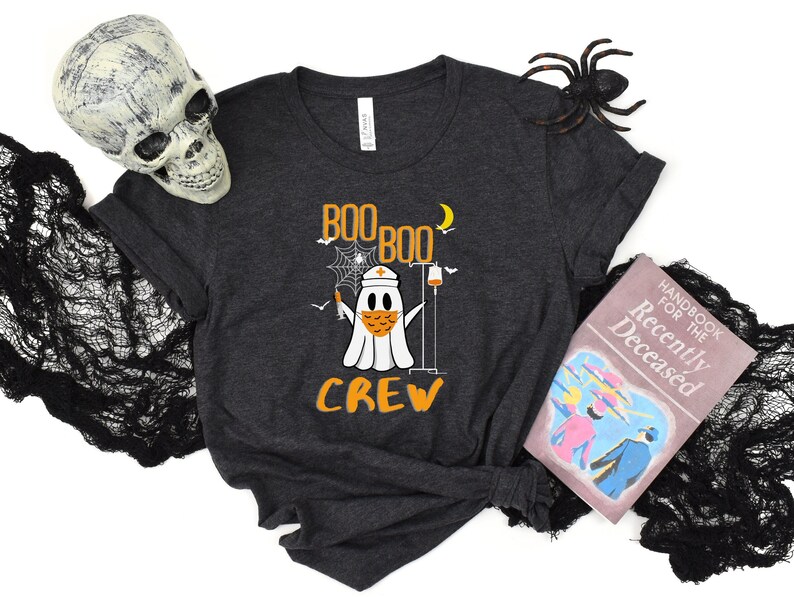 May include: A dark gray t-shirt with a ghost wearing a face mask and holding a syringe. The text "Boo Boo Crew" is printed on the shirt.
