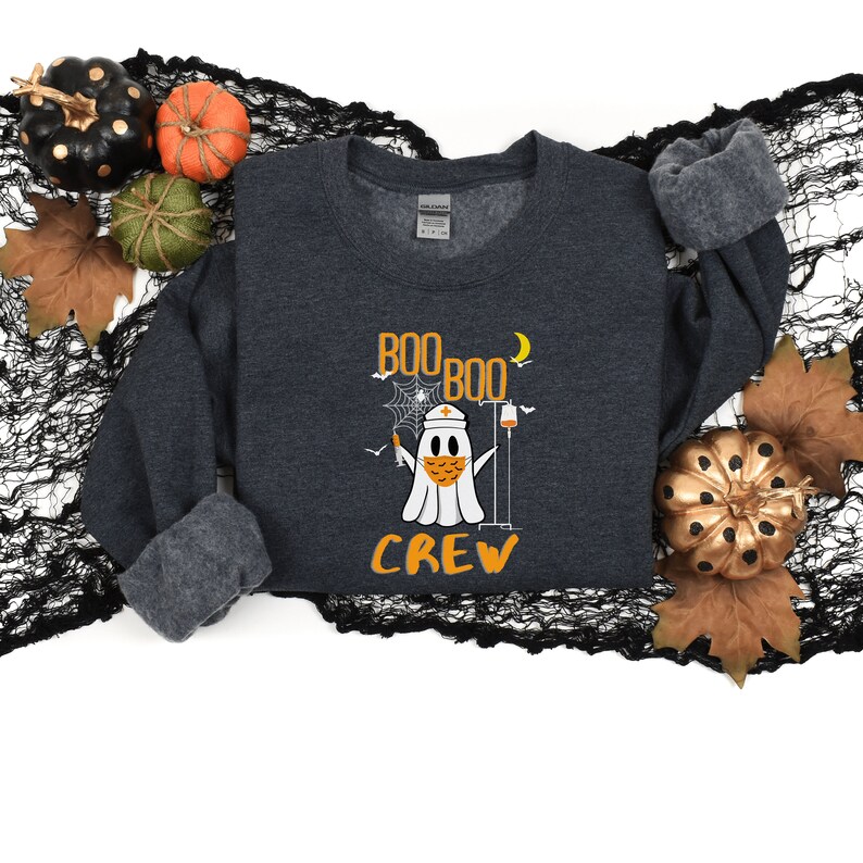 May include: A dark gray sweatshirt with the text "BOO BOO CREW" in orange letters. A cartoon ghost wearing a nurse's hat and a face mask is holding a syringe and a IV bag. The ghost is surrounded by a spiderweb and a crescent moon.