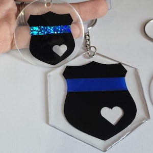 May include: Two keychains with a black and blue police badge design. One keychain has a blue glitter stripe and the other has a solid blue stripe. Both keychains have a white heart cutout.