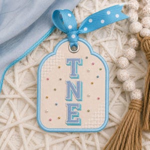 May include: A cream-colored fabric tag with a rounded top and blue stitched border, featuring the embroidered blue letters T, N, and E stacked vertically. The tag is decorated with small colorful beads and tied with a blue ribbon with white polka dots.