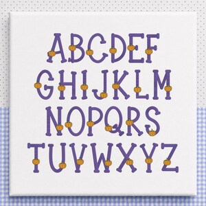 以下が含まれることがあります： 紫色の大文字で英語のアルファベットが描かれた白い正方形の看板。各文字には小さなオレンジ色のカボチャのイラストが装飾されています。文字は4行に配置されており、AからZまでの完全なシーケンスがはっきりと表示されています。