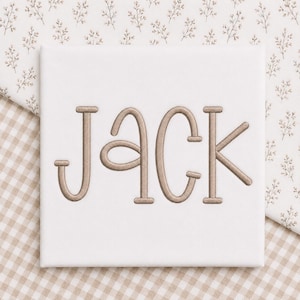May include: A square white fabric panel featuring the name JACK embroidered in a light brown thread. The letters are rendered in a stylized, rounded font with distinct horizontal bars on the J, A, C, and K characters, centered on the fabric.