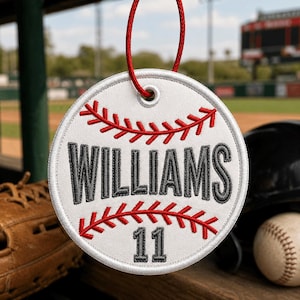 May include: A white baseball-themed ornament with red stitching and a red hanging cord. The name "WILLIAMS" and the number "11" are embroidered in gray. The ornament is round and has a baseball design.