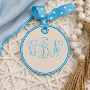 Op de afbeelding: Een rond beige ornament met een blauw geborduurd monogram met de letters CBN. Het ornament is afgewerkt met een blauwe stikselrand en bekroond met een blauw lint met witte stippen, vastgemaakt in een strik met een metalen oogje.