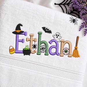 Puede incluir: Una toalla blanca con letras bordadas con temática de Halloween que deletrean el nombre Ethan. El diseño incluye un sombrero de bruja, un caldero burbujeante, una araña, una momia, un fantasma, un globo ocular y una escoba en tonos morados, verdes y naranjas.