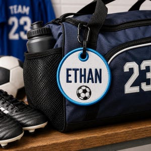 May include: A round white embroidered bag tag with a blue border, featuring the name ETHAN in dark blue capital letters and a small black and white soccer ball graphic, attached to a navy blue duffel bag with a black carabiner clip.