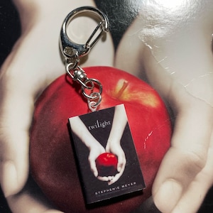 May include: A keychain with a miniature book cover that reads "twilight" and a red apple design. The keychain has a silver clasp. The background shows a red apple and a blurred image of hands.