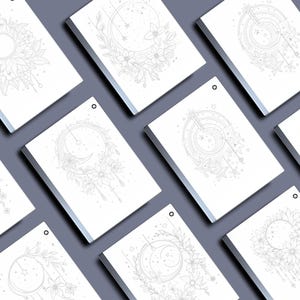 Peut inclure: Plusieurs cahiers blancs orn&eacute;s de dessins au trait sur le th&egrave;me c&eacute;leste. Chaque cahier pr&eacute;sente une illustration diff&eacute;rente de lunes, de soleils, d'&eacute;toiles et d'&eacute;l&eacute;ments floraux. Les cahiers sont dispos&eacute;s sur une surface grise.