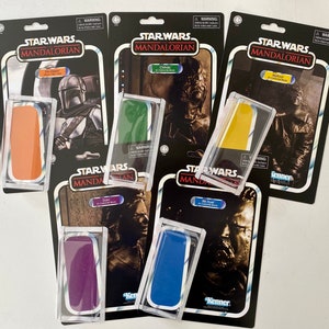 May include: A collection of eight Star Wars The Mandalorian action figure packaging. The packaging is in the style of vintage Kenner action figures. The packaging features different characters from the show, including The Mandalorian, Grogu, and Moff Gideon. The packaging is in various colors, including black, white, red, blue, green, and yellow.