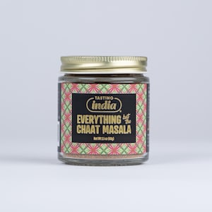 May include: A glass jar with a gold lid containing a brown spice blend. The label reads "Tasting India" in a gold circle with a black background. The label also reads "Everything but the Chaat Masala" in gold text on a black background. The jar is sitting on a white surface.
