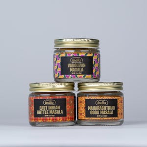 Puede incluir: Tres frascos de vidrio con tapas doradas y etiquetas coloridas que contienen diferentes mezclas de especias indias. Las etiquetas dicen "East Indian Bottle Masala", "Maharashtrian Goda Masala" y "Vadouvan Masala".