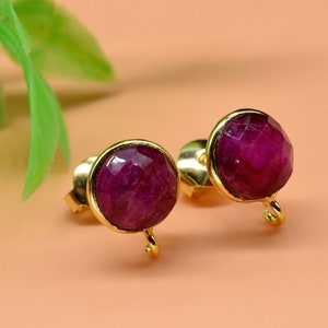 Pode incluir: Um par de brincos de pressão de ouro com uma pedra preciosa redonda, facetada, de cor rosa escura. Cada brinco tem um pequeno laço de ouro pendurado na parte inferior.