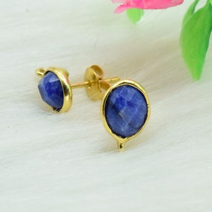 Könnte beinhalten: Ein Paar goldfarbene Ohrstecker mit ovalen, facettierten, tiefblauen Edelsteinen. Die Ohrringe sind in einer goldenen Lünette gefasst und werden auf einer weißen, strukturierten Oberfläche präsentiert.