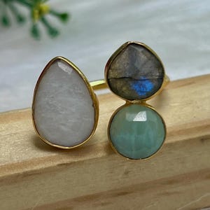 Könnte beinhalten: Ein goldener Ring mit drei tropfenförmigen Steinen. Die Steine sind weiß, grün und grau.