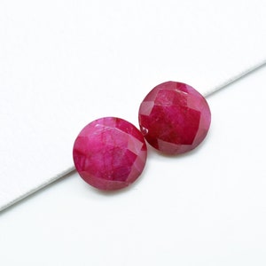 May include: Two round, faceted ruby gemstones, each with a deep red color, are displayed on a white surface.