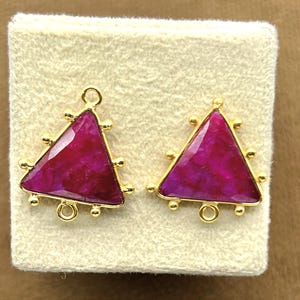 Peut inclure: Une paire de boucles d'oreilles dorées avec des pierres précieuses triangulaires de couleur magenta. Les boucles d'oreilles ont un cadre doré avec de petits accents ronds. Les pierres sont posées sur une surface crème.