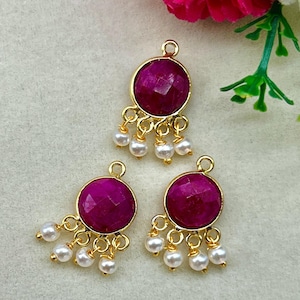 May include: Three gold-toned pendants with a round, faceted, magenta-colored stone and dangling white pearls. The pendants have a small loop at the top for hanging. The background is a light, neutral color.