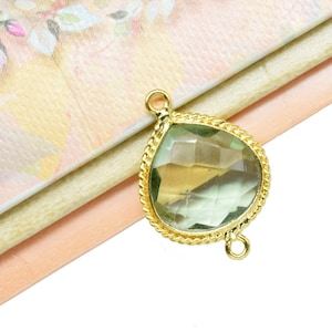 Op de afbeelding: Goudkleurige traanvormige hanger met een groene edelsteen. De hanger heeft twee lussen om aan een ketting of koord te bevestigen.