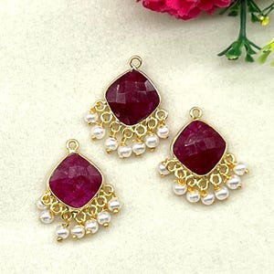 May include: Three gold-toned pendants with a square, faceted, deep red gemstone center. Each pendant has a fringe of small, white, round pearls. The pendants are displayed on a light-colored surface, with pink flowers in the background.