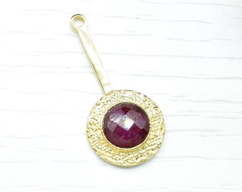 Op de afbeelding: Een gouden hanger met een rond, getextureerd frame en een donkerrode edelsteen in het midden.
