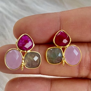 Pode incluir: Um par de brincos de ouro com três pedras cada. As pedras são rosa, cinza e um rosa mais escuro.