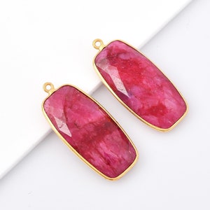 Può includere: Due pietre preziose rettangolari, sfaccettate, color rubino, con castoni dorati. Ogni pietra ha un piccolo anello nella parte superiore per il fissaggio a una catena o ad un altro componente di gioielleria. Le pietre sono su sfondo bianco.