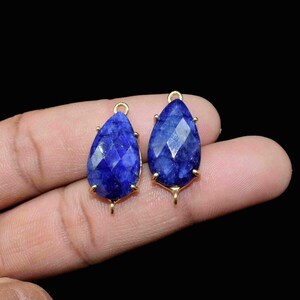 Pode incluir: Dois pingentes de pedra preciosa azul em forma de lágrima com engastes de metal dourado. Os pingentes têm um design facetado e são presos a um laço dourado.