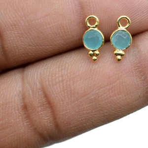 Puede incluir: Dos abalorios de pendientes de color dorado con una piedra redonda de color azul claro. Cada abalorio tiene tres pequeñas cuentas doradas que cuelgan de la parte inferior.