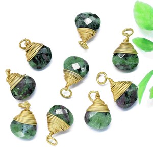 May include: Nine green and black gemstone pendants wrapped in gold wire. The pendants are arranged on a white surface with a green leafy branch in the background.