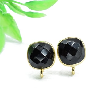 May include: A pair of square-shaped black gemstone stud earrings with a gold-colored bezel and a small loop for attachment. The gemstones have a faceted cut, reflecting light. The earrings are set against a white background.