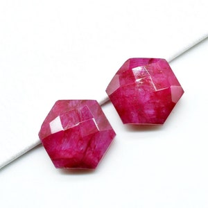 May include: Two faceted, pink ruby gemstones, each with a hexagonal shape, are displayed on a white surface.