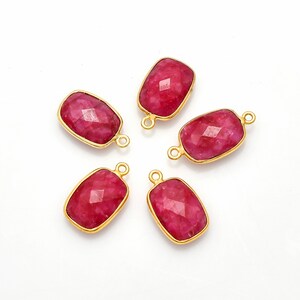 May include: Five rectangular ruby gemstones with gold-colored bezels. Each gemstone has a faceted surface and a small loop at each end. The gemstones are arranged on a white surface.