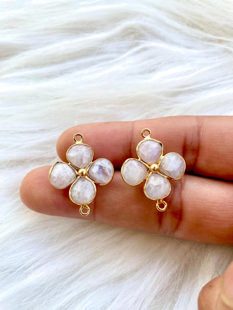 May include: Two gold-tone flower-shaped charms with a white gemstone center. Each charm has four petals and a loop for attaching to jewelry.