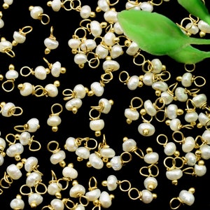 Puede incluir: Una colecci&oacute;n de abalorios de perlas doradas con un bucle en la parte superior. Los abalorios est&aacute;n esparcidos sobre una superficie negra con una hoja verde en el fondo.
