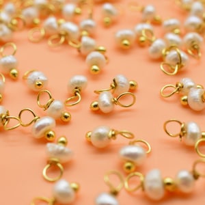 Puede incluir: Una colecci&oacute;n de abalorios de perlas blancas y doradas, cada uno con un peque&ntilde;o bucle para sujetar a joyas u otras manualidades. Los abalorios est&aacute;n esparcidos sobre una superficie de color naranja claro.