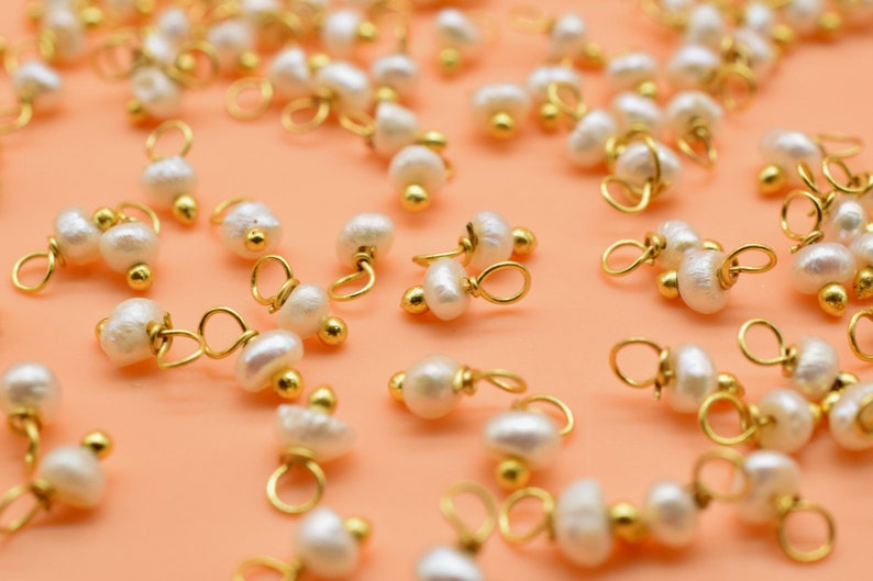 Puede incluir: Una colecci&oacute;n de abalorios de perlas blancas y doradas, cada uno con un peque&ntilde;o bucle para sujetar a joyas u otras manualidades. Los abalorios est&aacute;n esparcidos sobre una superficie de color naranja claro.