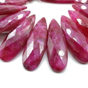 May include: A close-up of a strand of faceted ruby gemstone beads. The beads are a deep pink color and have a pear shape. The beads are arranged in a semi-circle.
