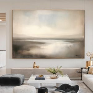 Könnte beinhalten: Ein großes, rechteckiges abstraktes Landschaftsgemälde mit goldenem Rahmen. Das Kunstwerk zeigt eine gedämpfte Farbpalette aus Beige, Grau und Hellblau, die eine ruhige, atmosphärische Szene darstellt. Das Gemälde hängt über einem modernen Wohnzimmer.