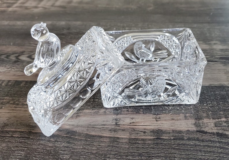 May include: A clear, rectangular glass trinket box with a lid. The lid features a small bird figurine and the box is decorated with etched bird and floral designs. The box is open, revealing the interior. The glass has a cut crystal appearance.