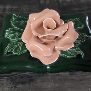 May include: A ceramic decorative piece featuring a large, light pink rose atop a dark green rectangular base. The base has embossed leaves with green accents. The rose has detailed petals and a glossy finish.
