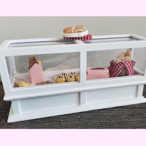 May include: White display case with glass shelves, showcasing miniature baked goods, including pastries, bread, and a loaf of bread in a red and white checkered basket.