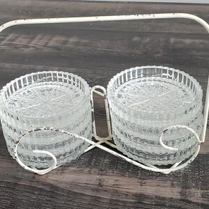 May include: A white metal stand with two stacks of clear glass coasters. The coasters are round and have a textured surface.